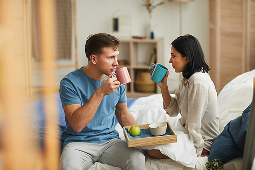 Warm-toned portrait of happy young couple enjoying breakfast in bed and looking at each other in cozy home interior, copy space