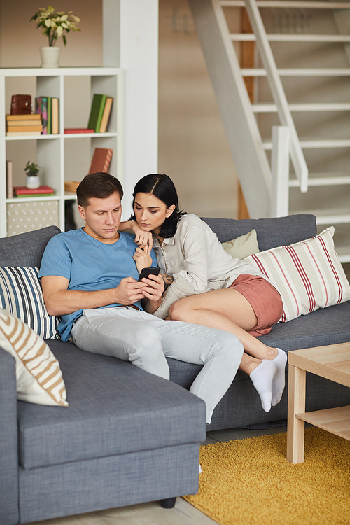 Vertical full length portrait of modern young couple using smartphone while relaxing on sofa in cozy home interior, copy space