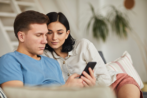 Portrait of modern young couple using smartphone while relaxing on sofa in cozy home interior, copy space