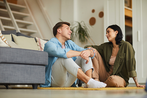 Warm toned full length portrait of modern young couple talking to each other sincerely while sitting on floor in cozy home interior, copy space