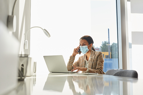 Portrait of mixed-race businesswoman wearing mask and speaking by smartphone while working in white office, copy space