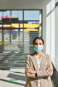 Vertical waist up portrait of confident businesswoman wearing mask while standing with arms crossed in sunlight, copy space