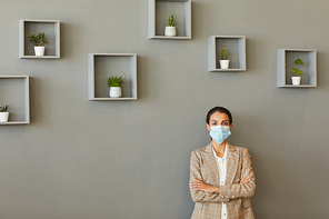 Minimal waist up portrait of confident businesswoman wearing mask while standing with arms crossed against grey wall, copy space