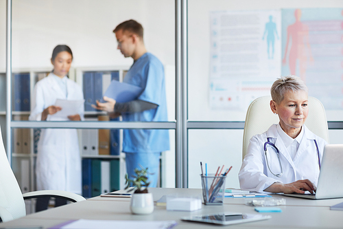 Portrait of mature female doctor using laptop while working in office with other people in background, copy space