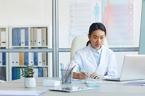 Portrait of female Asian doctor using laptop while sitting at workplace in medical office interior, copy space