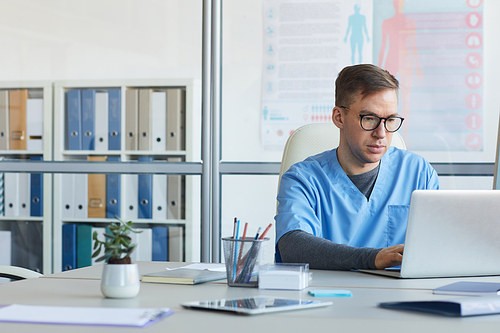 Portrait of male doctor using laptop and wearing glasses while sitting at workplace in medical office interior, copy space