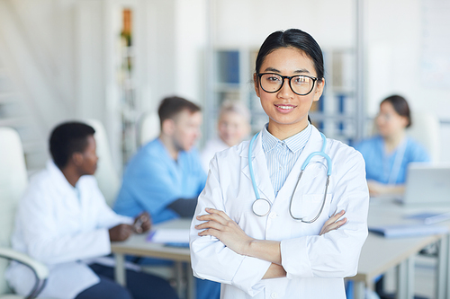 Waist up portrait of female Asian doctor standing with arms crossed and smiling at camera against medical conference background, copy space