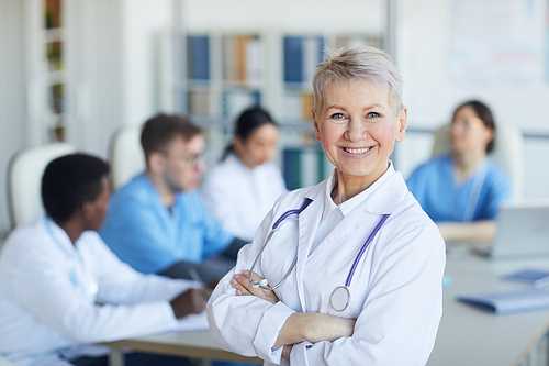 Waist up portrait of senior female doctor standing with arms crossed and smiling at camera against medical conference background, copy space