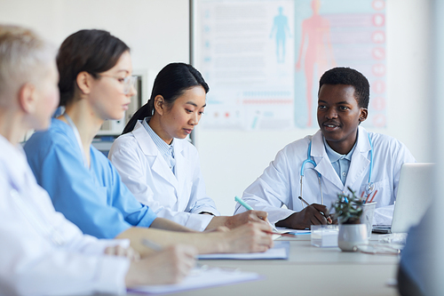 Multi-ethnic group of young doctors sitting round table during medical council or conference in clinic, copy space