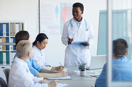 Portrait of young African-American doctor giving speech while standing at table during medical council or conference in clinic, copy space