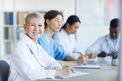 Portrait of mature female doctor smiling at camera while sitting at table during medical council or conference in clinic, copy space