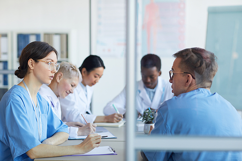 Portrait of young female doctor talking to colleague while sitting at table during medical council or conference in clinic, copy space