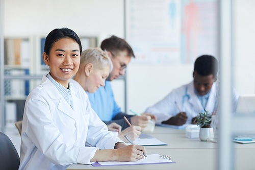 Portrait of Asian female doctor smiling at camera while sitting at table during medical council or conference in clinic, copy space