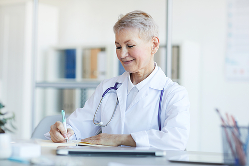 Portrait of mature female doctor writing notes and smiling while sitting at desk in clinic interior