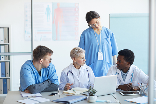 Multi-ethnic group of doctors communicating while sitting at desk with laptop in clinic office interior, copy space