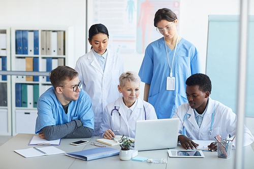 Multi-ethnic group of doctors using laptop together while sitting at desk in medical office interior, copy space