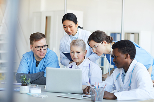 Multi-ethnic group of doctors using laptop together while sitting at desk in medical office interior