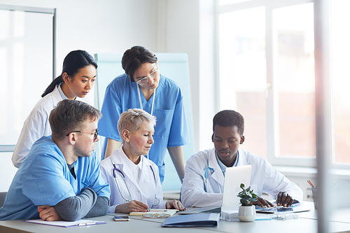 Multi-ethnic group of doctors looking at laptop screen while working together sitting at desk in medical office interior