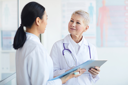 Waist up portrait of mature female doctor talking to trainee while standing in medical office interior, copy space