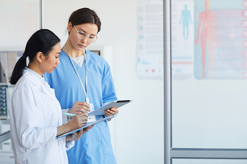 Side view portrait of two female medics filling forms while standing in medical office interior, copy space