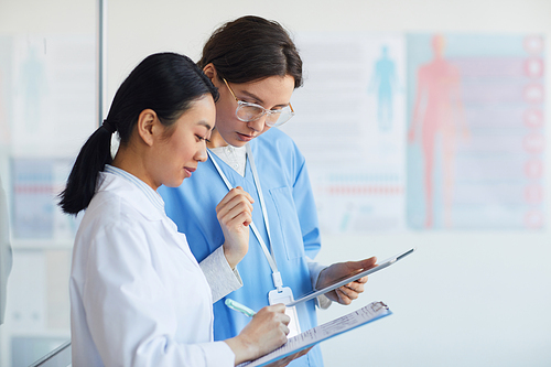 Side view portrait of two female medics writing on clipboard while standing in medical office interior, copy space