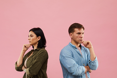 Waist up portrait of pensive young couple standing back to back against pink background, copy space