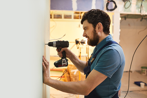 Side view portrait of bearded construction worker drilling wall while renovating house alone, copy space