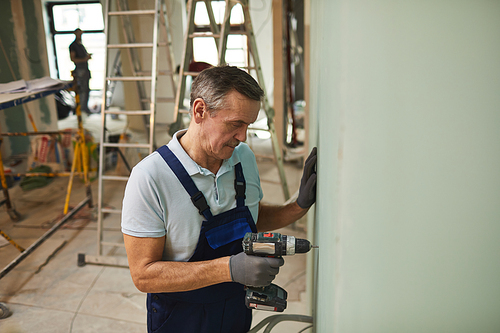 Portrait of senior construction worker drilling wall while renovating house alone, copy space