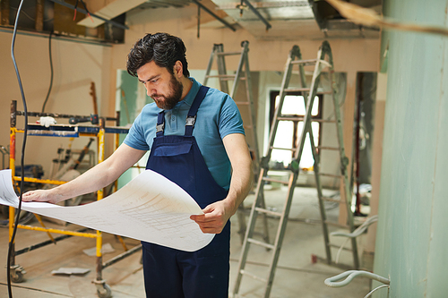 Portrait of bearded construction worker looking at floor plans while renovating house alone, copy space