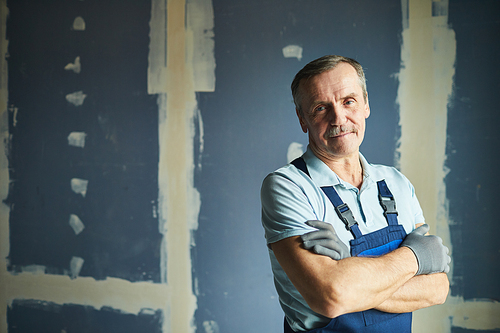 Waist up portrait of senior construction worker smiling at camera while posing confidently against dry wall, copy space