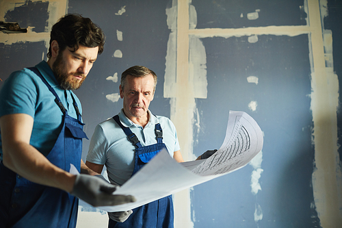 Portrait of two construction workers holding plans and standing against dry wall while renovating house, copy space