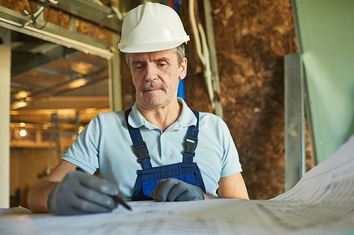 Waist up portrait of senior construction worker wearing hardhat while looking at floor plans while renovating house, copy space