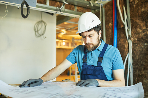 Portrait of bearded construction worker wearing hardhat and looking at floor plans while renovating house, copy space