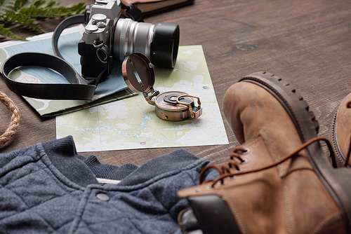 Close-up of compass and photographic camera on maps placed on wooden table with boots and sweater, flat lay