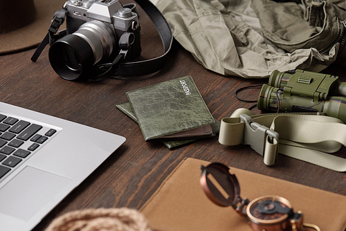 Close-up of passports in green covers, belt, binoculars, photographic camera, jackets and laptop on wooden table