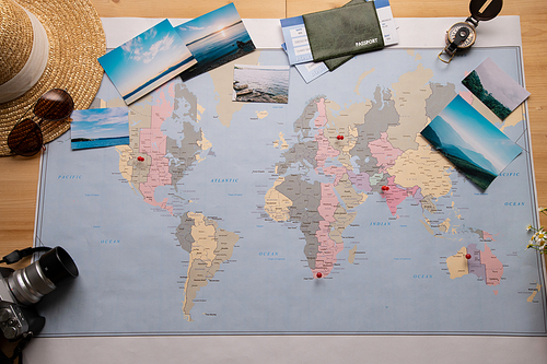 Above view of paper world map with red pins and beautiful nature photographs, camera, compass, sun hat and passports around