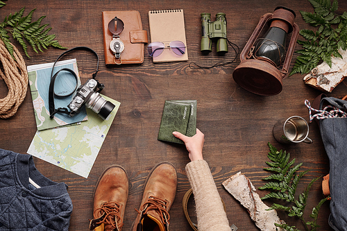 Above view of woman taking passports in green covers from wooden table with hiking stuff while preparing for journey