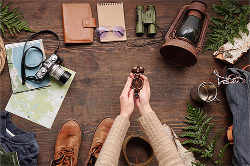 Unrecognizable hiker in sweater holding open compass above wooden table with hiking stuff and green twigs