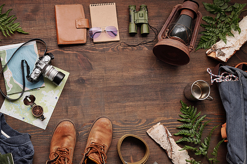 Above view of lantern, binoculars, sketchpad, purse, camera, maps, boots, belt and green twigs in circle on wooden table