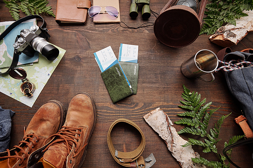 Above view of prepared tickets and passports for foreign adventure, green twigs, boots, belt, camera and map on table, flat lay