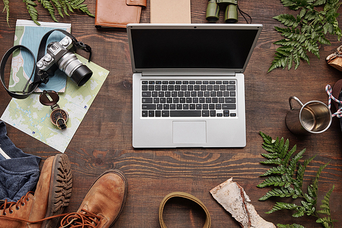 Flat lay composition symbolizing active tourism: laptop, maps, thermos mug, belt, compass on wooden table