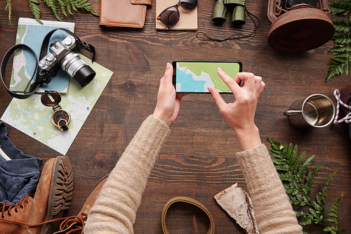 Above view of woman checking hiking route on online map while using smartphone above wooden table with stuff, flat lay