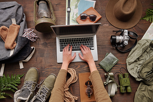 Female hands typing on laptop at table with hiking stuff such as clothes, rope, lantern, maps, flat lay