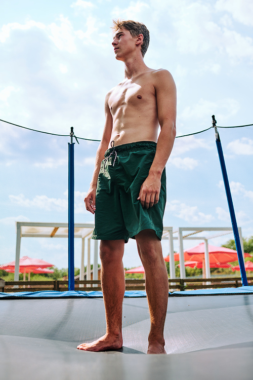 Young topless athlete in green shorts standing on trampoline surrounded by net while going to jump in natural environment over cloudy sky
