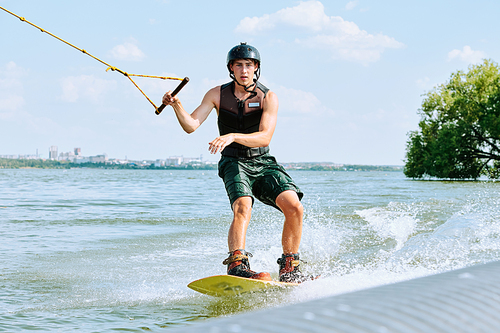 Young sportsman in shorts, safety jacket and helmet holding by handle with rope while moving on surfboard during outdoor training