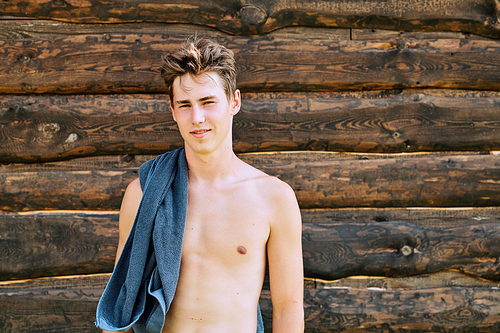 Young topless sportsman with blue towel on shoulder standing against wooden wall in front of camera after outdoor sports training