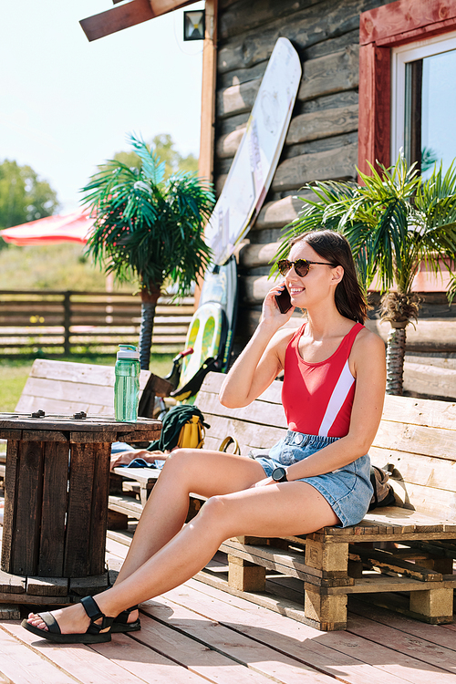 Happy young brunette female in sunglasses, red tanktop and denim shorts sitting on wooden bench outdoors and talking on smartphone