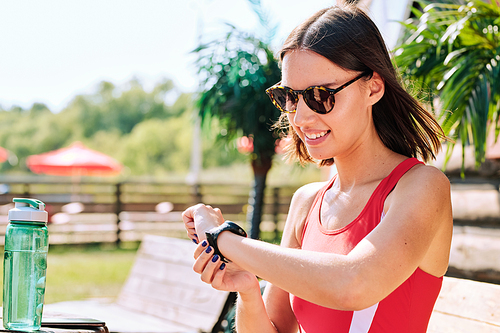 Smiling young brunette female in sunglasses and red tanktop looking at wristwatch and having water on hot summer day at resort