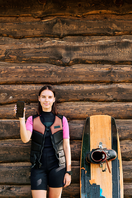 Young female surfer in safety jacket and sportswear standing against wall of wooden house and showing her selfie in smartphone