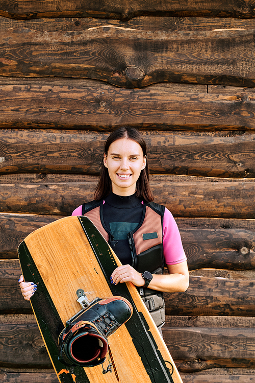 Active young female surfer in safety jacket holding surfboard while standing against wall of wooden house in front of camera